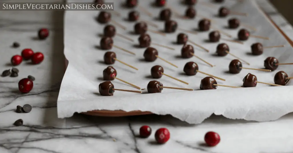 chocolate covered cranberries cooling on parchment paper on cookie sheet on marble table with loose cranberries and chocolate chips