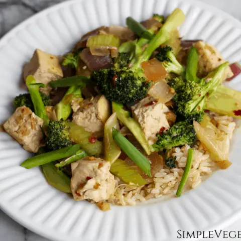 Sichuan broccoli and tofu with celery, green and red onions, and brown rice served on white plate on white marble table