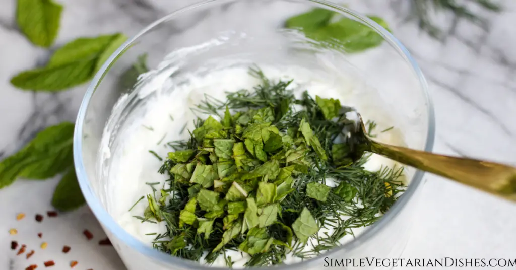 fresh chopped dill and mint on top of haydari yogurt base in glass bowl with gold fork with mint in background