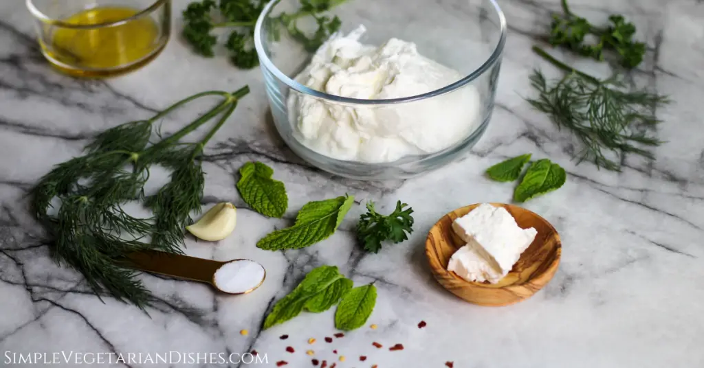 ingredients for haydari greek yogurt feta mint garlic dill parsley red pepper flakes and olive oil on white marble table