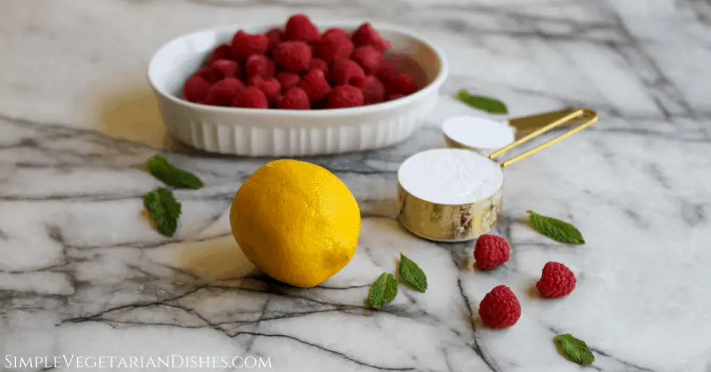 Melba sauce ingredients on marble table raspberries in white dish, lemon, powdered sugar and cornstarch in gold measuring cups and spoons, fresh mint