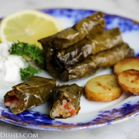 dawali stuffed grape leaves served with roasted potatoes, greek yogurt garnished with lemon slice and parsley on blue china on white marble table