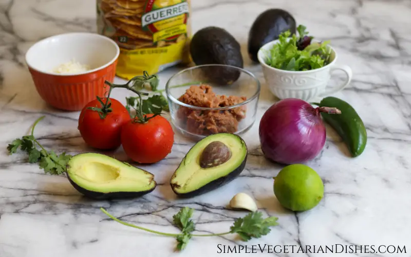 tostaguac ingredients on white marble table avocados, tomatoes, refried beans, cheese, onion, tostada shells, garlic, jalapeño, lettuce, cilantro