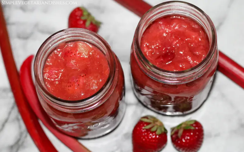 two mason jars of strawberry rhubarb sauce with fruit in background