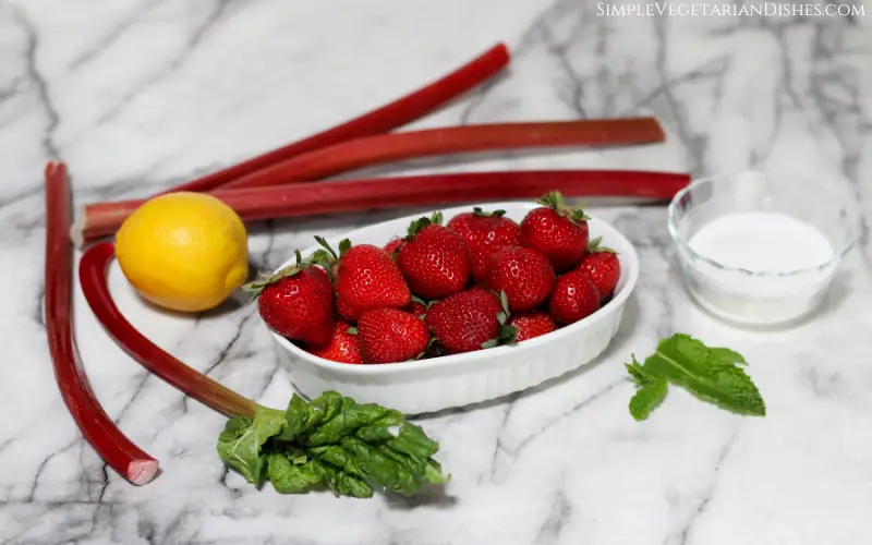 dish of strawberries with rhubarb stalks, lemon, mint, and sugar in background