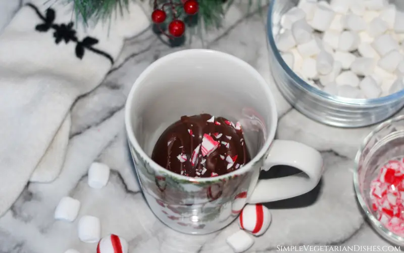 vegan hot chocolate bomb in holiday mug with peppermint candies and vegan marshmallows in background